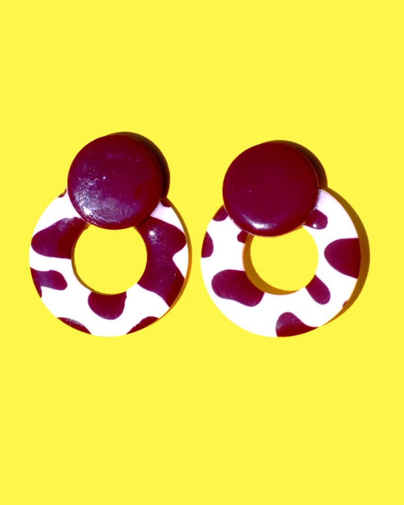 Paire de boucles d'oreilles en forme de cercle ajouré. Une grosse puce ronde en relief est collée sur le haut de la boucle. La boucle est au motif tâches de vache brunes sur fond blanc. La "puce" ronde est marron unie.