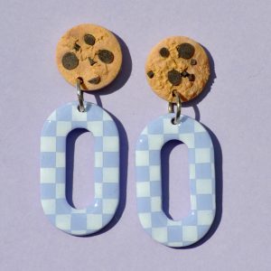 Paire de boucles d'oreilles en forme d'ovale. La paire de boucles d'oreilles est ajourée au centre. Une puce cookie surmonte une boucle motif damier blanc sur fond violet pâle.