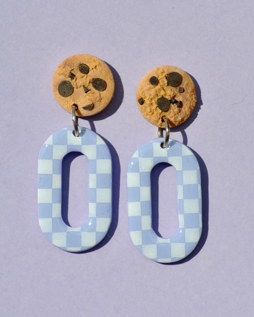 Paire de boucles d'oreilles en forme d'ovale. La paire de boucles d'oreilles est ajourée au centre. Une puce cookie surmonte une boucle motif damier blanc sur fond violet pâle.