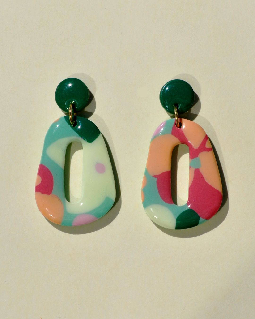 Paire de boucles d'oreilles en forme galet donut. La paire de boucles d'oreilles est ajourée au centre d'un long rectangle arrondi élancé. Une petite puce ronde verte foncée unie surmonte une boucle au motif floral printanier.