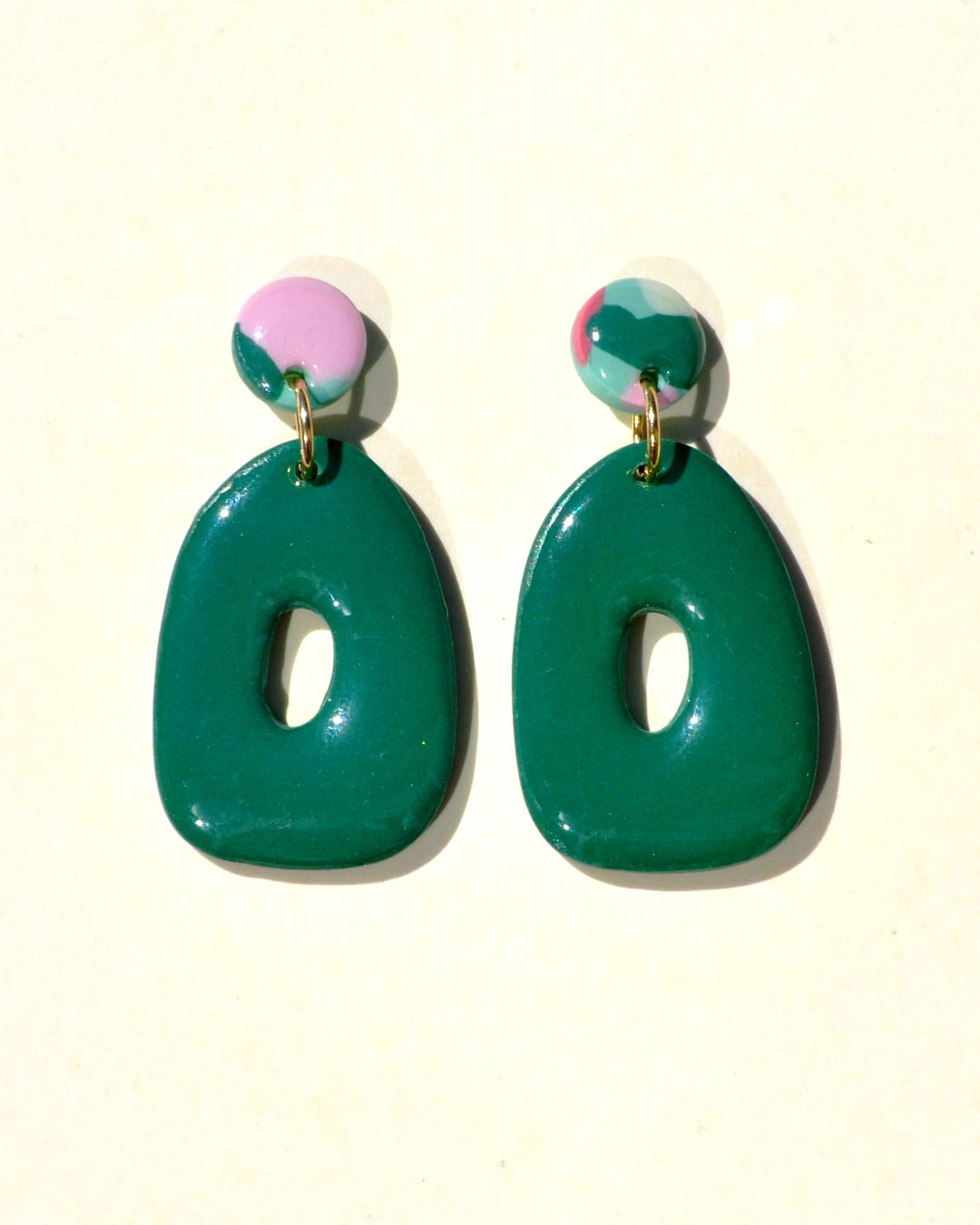 Petite paire de boucles d'oreilles en forme petit galet donut. La paire de boucles d'oreilles est ajourée au centre d'un petit rond abstrait. Une petite puce ronde motif floral printanier surmonte une boucle unie vert foncée.