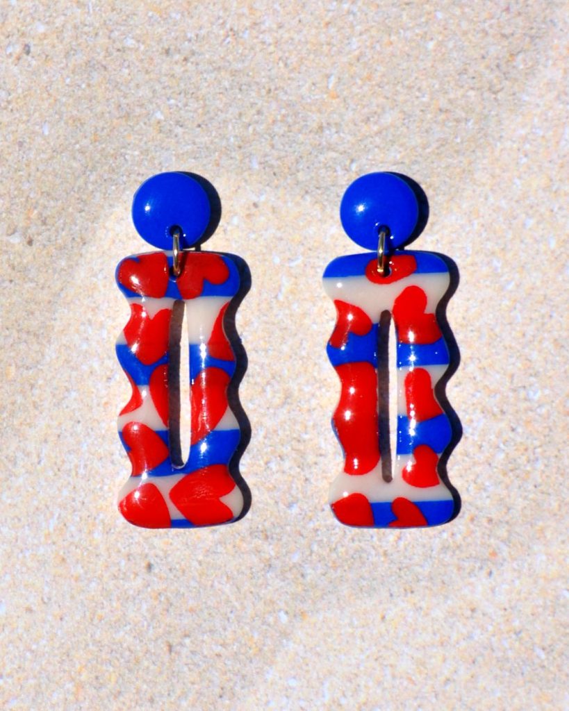 Paire de boucles d'oreilles en forme "petit lu" festonné allongé. La paire de boucles d'oreilles est ajourée au centre d'une ellipse très fine. Une petite puce unie bleu éclatant surmonte une boucle motif marinière cœurs rouges.