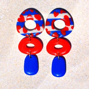 Paire de boucles d'oreilles abstraite en trois parties. La puce est la partie la plus imposante de la boucle en triangle dodu arrondi ajouré, au motif marinière cœurs rouges. La deuxième partie de la boucle est un ovale horizontal ajouré rouge vif. Une pampille rectangle arrondi vertical bleu éclatant vient terminer la boucle.