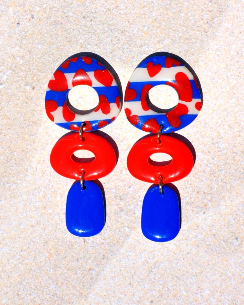 Paire de boucles d'oreilles abstraite en trois parties. La puce est la partie la plus imposante de la boucle en triangle dodu arrondi ajouré, au motif marinière cœurs rouges. La deuxième partie de la boucle est un ovale horizontal ajouré rouge vif. Une pampille rectangle arrondi vertical bleu éclatant vient terminer la boucle.