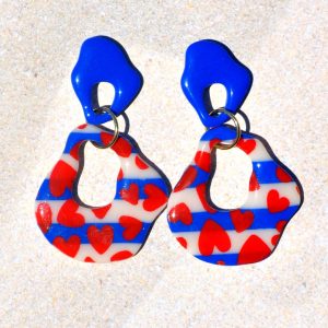 Paire de boucles d'oreilles duo de formes abstraites dodues et ajourés. Une petite puce abstraite bleu éclatant surmonte une plus grosse forme abstraite marinière à cœurs rouges.