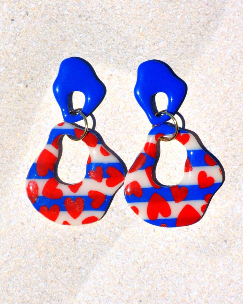 Paire de boucles d'oreilles duo de formes abstraites dodues et ajourés. Une petite puce abstraite bleu éclatant surmonte une plus grosse forme abstraite marinière à cœurs rouges.