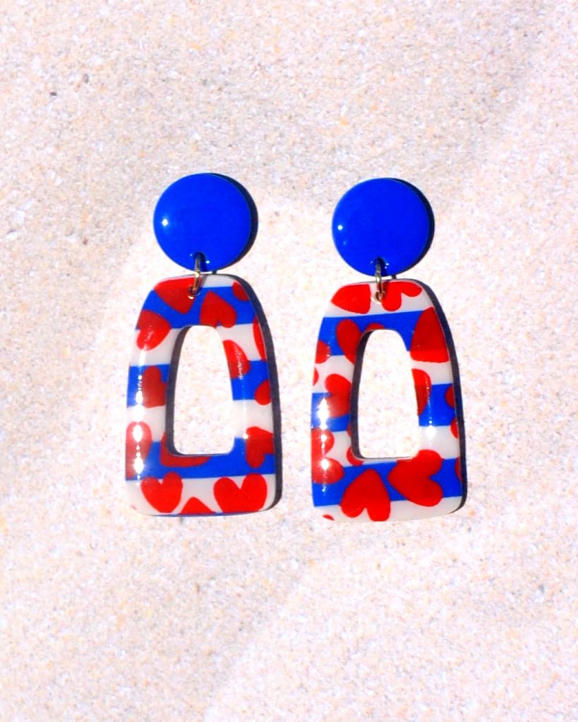 Paire de boucles d'oreilles en deux parties. Une puce ronde unie bleue éclatante surmonte un quadrilatère trapézoïde ajouré, au motif marinière cœurs rouges.
